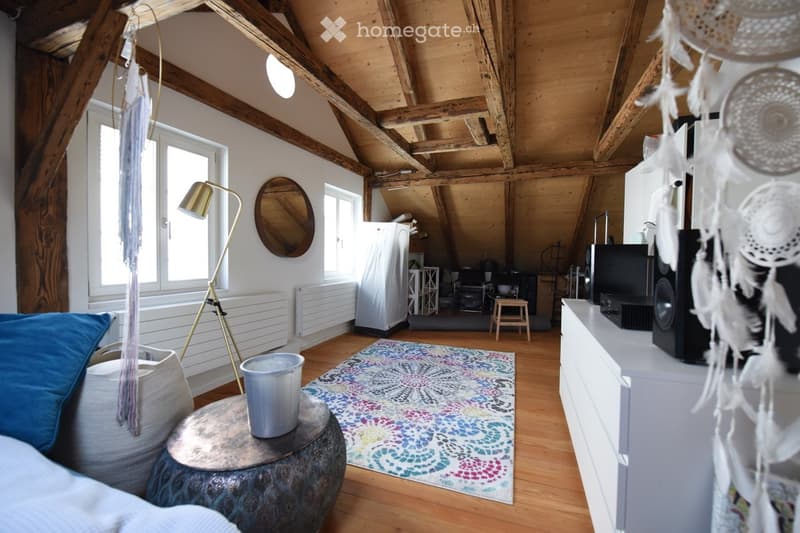A cozy, loft-style living space with exposed wooden beams, a large window, a patterned rug, a sofa, a small table with a bucket, a floor lamp, a mirror, a refrigerator, and a television set.