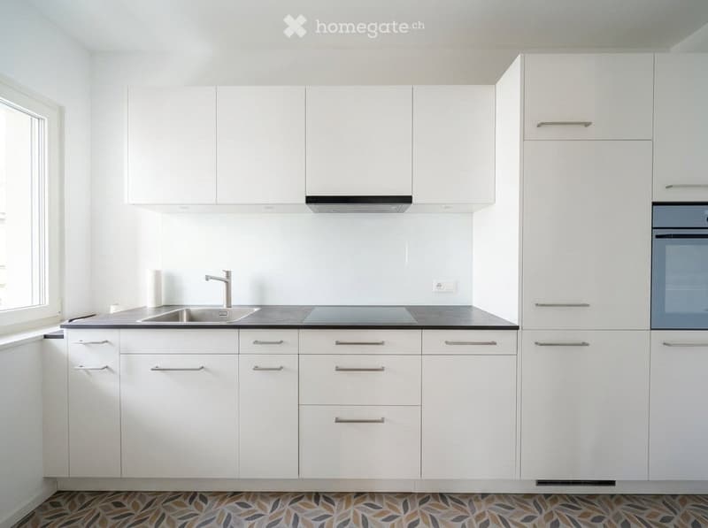 Modern white kitchen with sleek cabinets, stainless steel sink and faucet, built-in oven, and a decorative patterned tile floor.