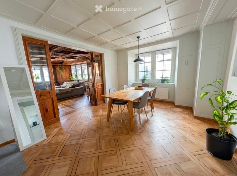 Bright dining area with wooden herringbone flooring, a large wooden dining table surrounded by modern chairs, a cozy sofa in the adjacent living space, and a large mirror on the wall.