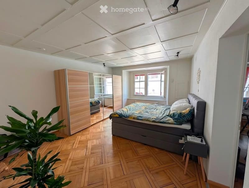 Modern bedroom with wooden flooring, large window, grey upholstered bed, nightstand, wardrobe, and a leafy plant in the corner.