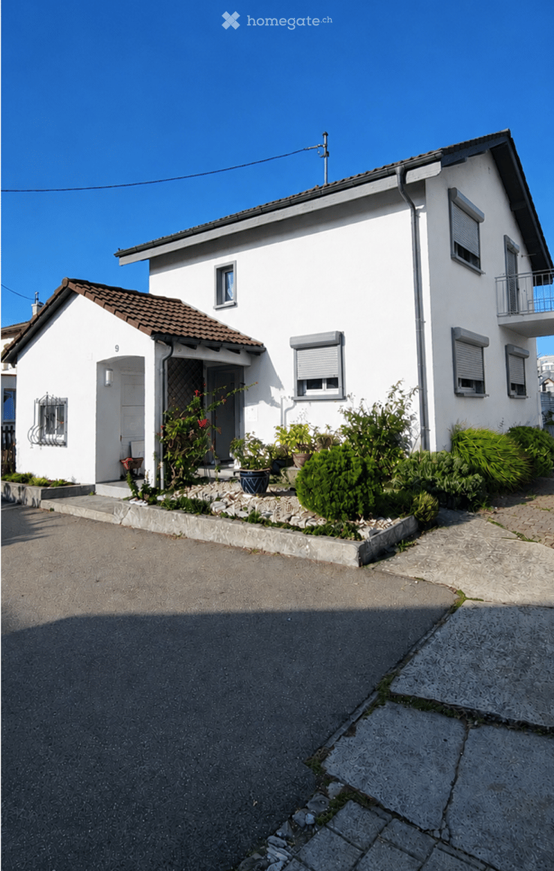 Two-story white house with a tiled roof, balcony, and small garden area with potted plants.