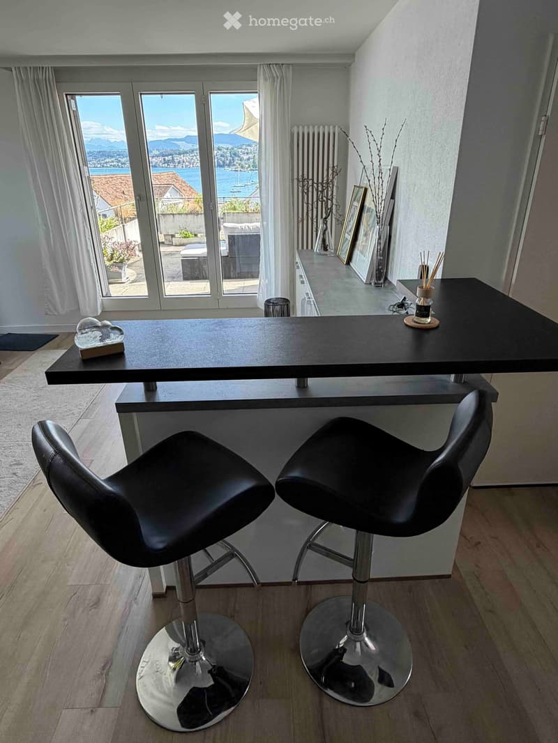 Modern room with wooden flooring, large windows offering a scenic view, a black table with two black stools, framed pictures, and a vase on a shelf.