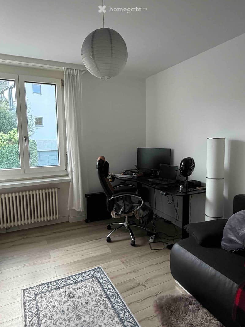 A modern, multi-functional room with a wooden floor, a large window with white curtains, a black sofa, a desk with a laptop and monitor, an office chair, and a decorative rug.