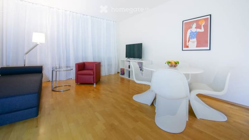 A modern, well-lit living room with a dark blue sofa, red armchair, white dining chairs, a small table, a television on a stand, and a floor lamp. The room features wooden flooring and white walls with a decorative picture frame above the TV.