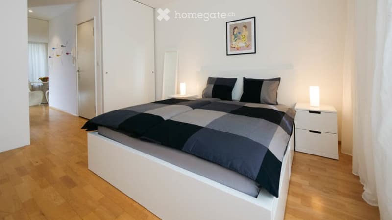 Modern bedroom with a large bed, nightstands, lamps, and a mirror. The room features light wooden flooring and white walls.