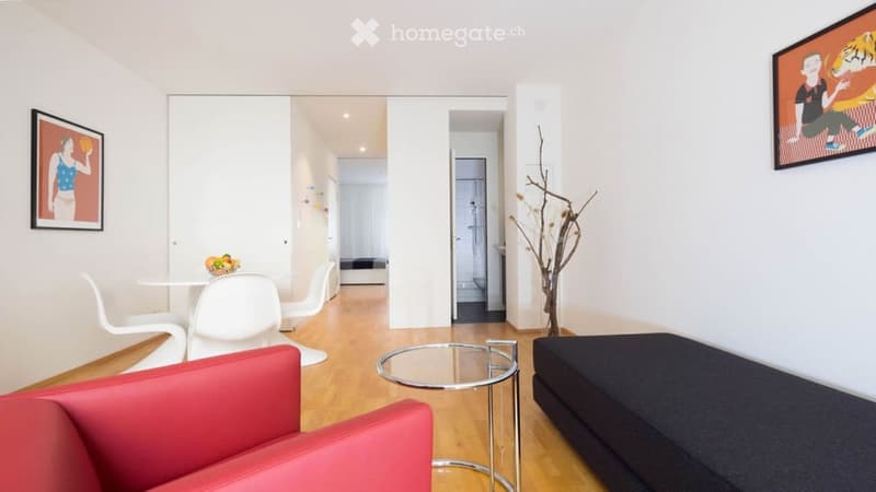Modern living room with a red sofa, glass coffee table, white chairs, and a small dining area visible in the background.
