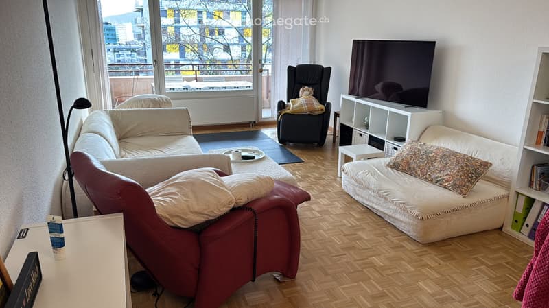 Modern living room with wooden flooring, large windows offering a city view, a red leather sofa, a white sofa, a black armchair, a small table, and a TV on a shelf.