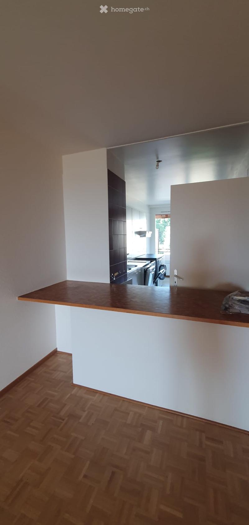 Empty room with wooden flooring, white walls, and a wooden countertop leading to a kitchen area with cabinets and appliances.
