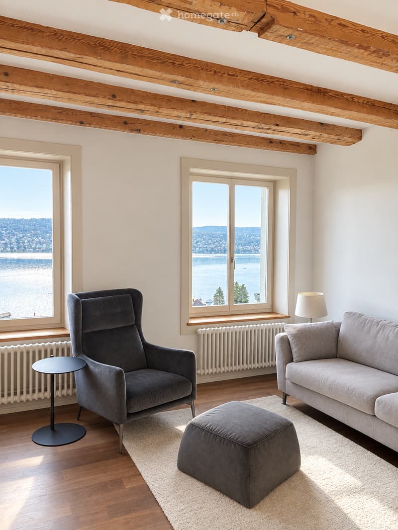 Modern living room with exposed wooden beams, large windows offering a scenic view, grey sofa, armchair, ottoman, small round table, and a floor lamp.