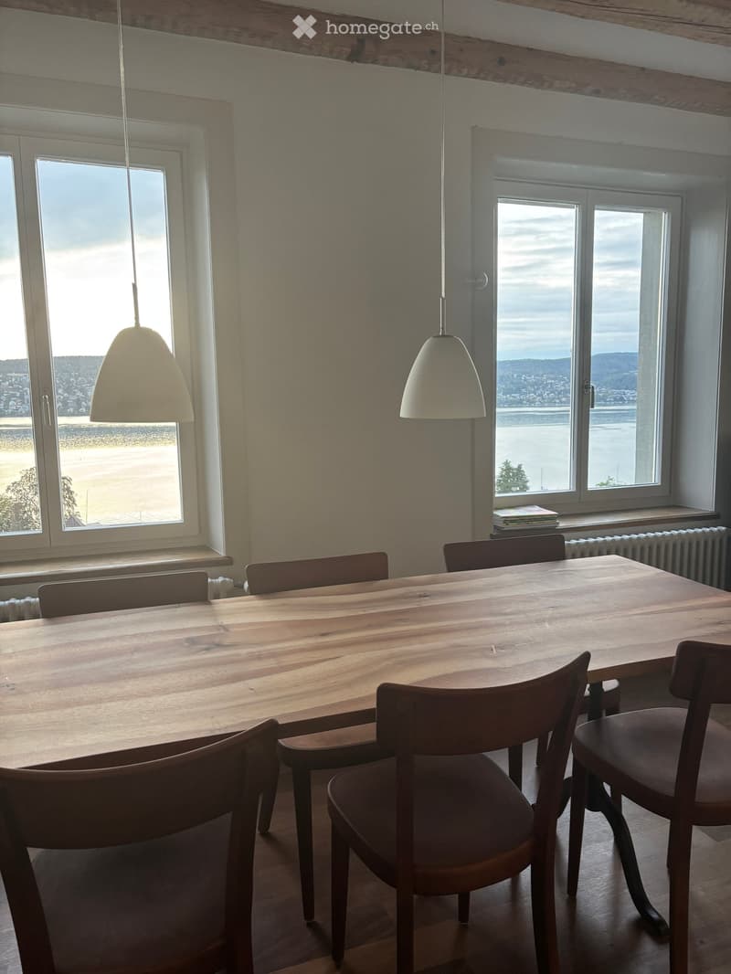 Bright dining area with wooden table and chairs, large windows offering a scenic view of the sea and distant hills.