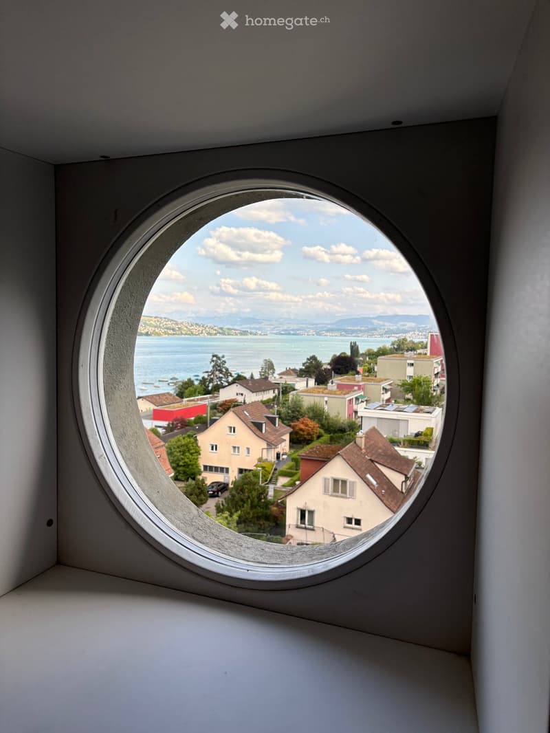 A round window with a scenic view of a lake, distant mountains, and surrounding houses.