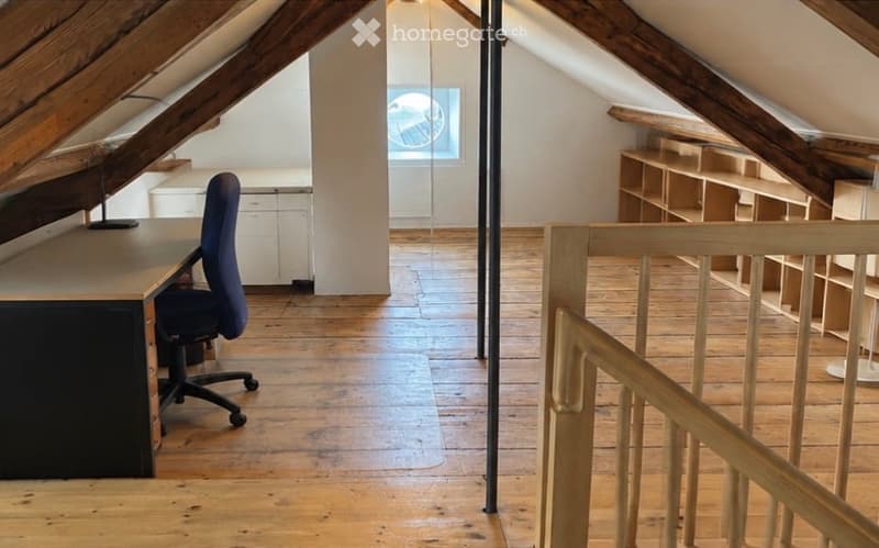 Spacious attic room with wooden floors, exposed wooden beams, a desk with a chair, built-in shelves, and a skylight window.