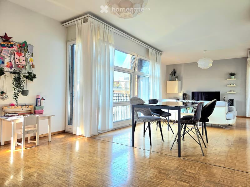 Bright living and dining area with wooden flooring, large windows, and modern furniture including a sofa, dining table, chairs, desk, and shelves.