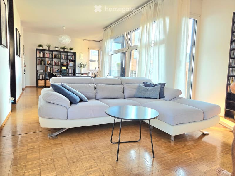 Modern living room with a grey sectional sofa, coffee table, bookshelf, and large windows with white curtains.