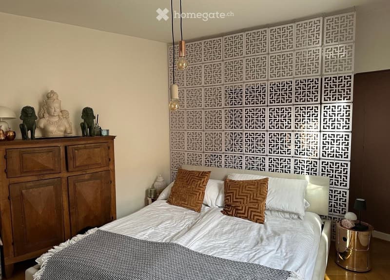 A modern bedroom with a large bed, patterned pillows, a wooden cabinet, and decorative statues on top. The walls feature a geometric tile design, and there are table lamps on either side of the bed.