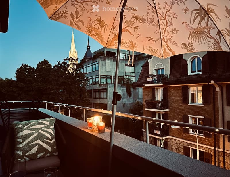Balcony with a sofa, decorative pillows, candles, and an umbrella, overlooking a cityscape with a church spire.