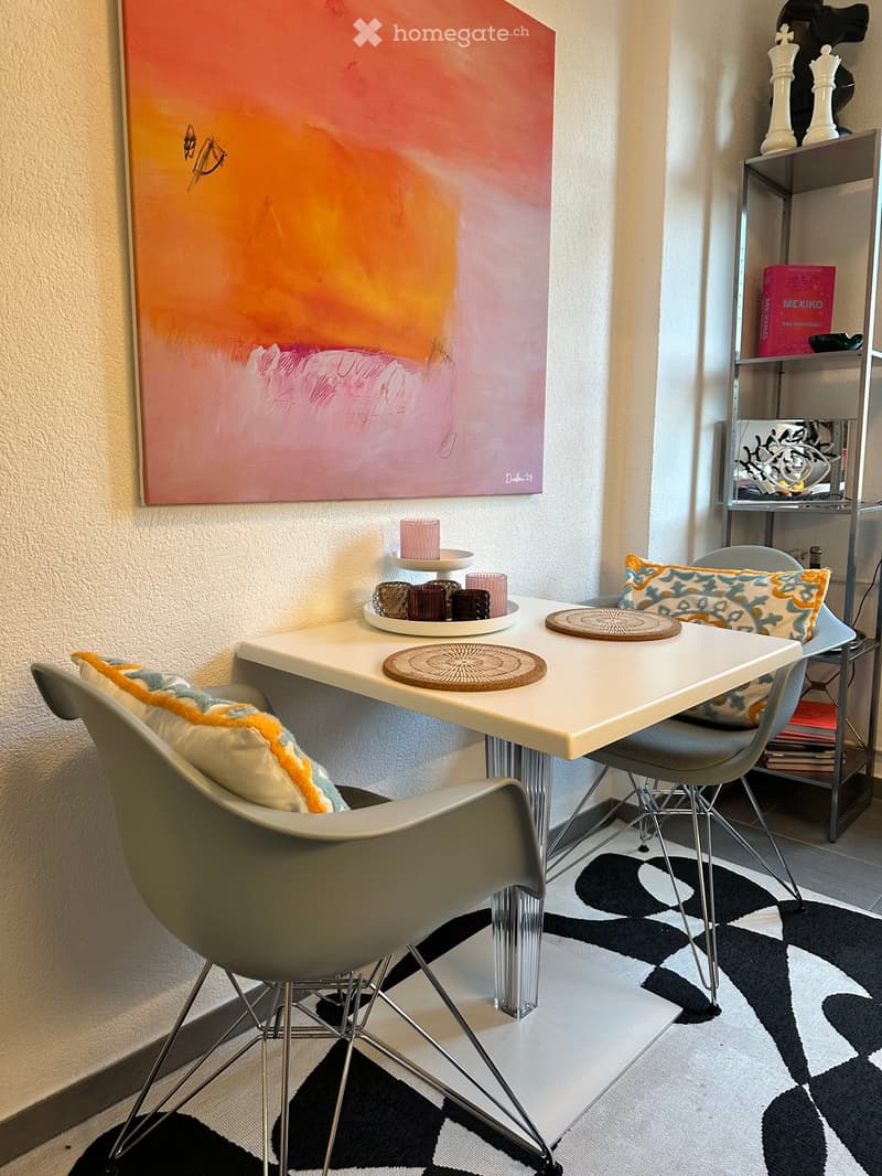 A modern, minimalist room with a white table, two chairs, a shelf with books and decorative items, and a large abstract painting on the wall.