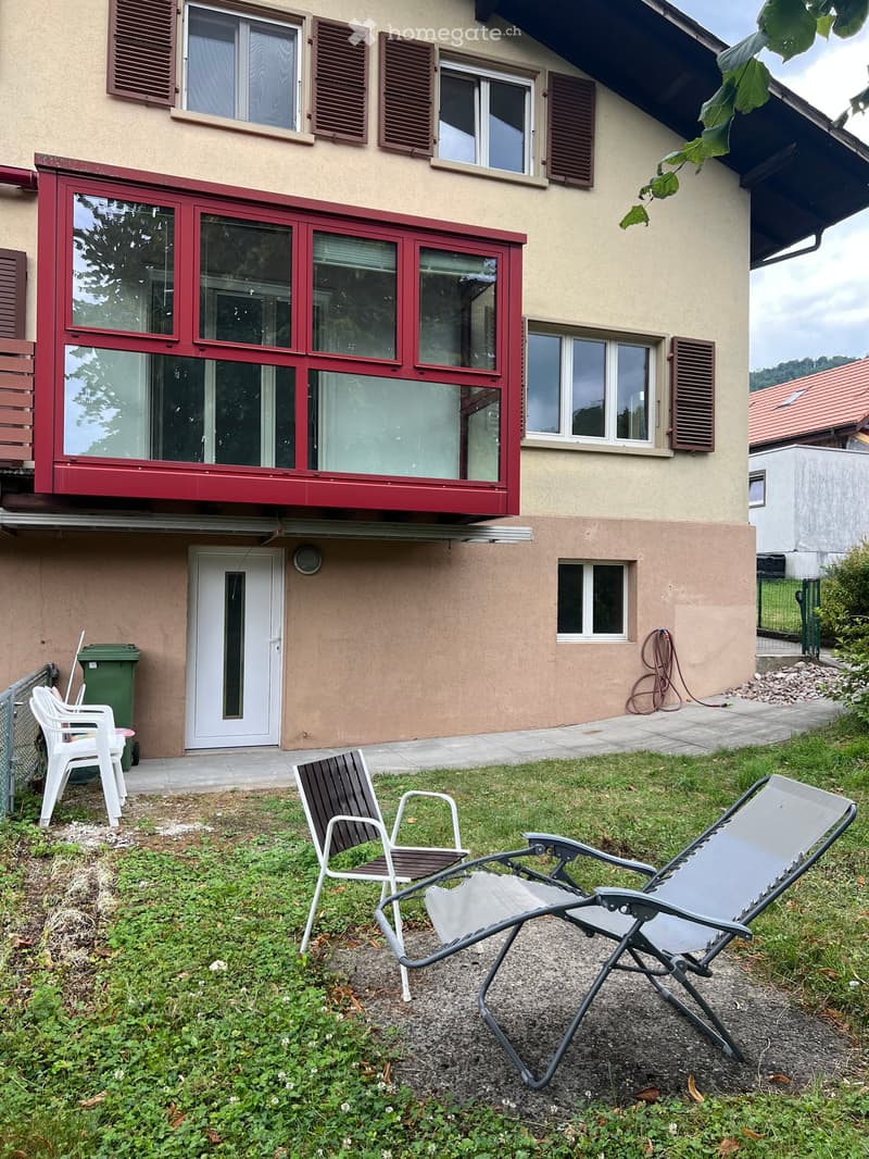 Garden area with a house in the background, featuring several chairs and a green trash bin near the entrance.
