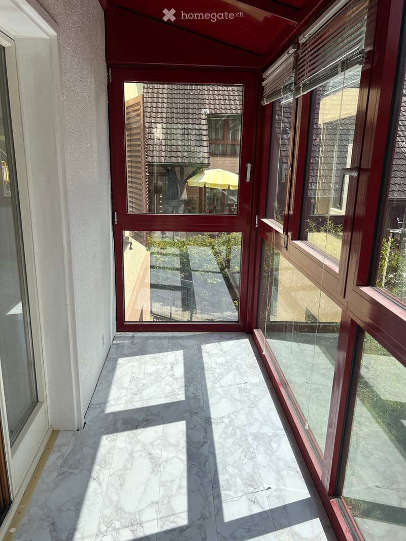 Marble-tiled balcony with large windows and sliding doors, offering views of a neighboring roof and garden area.