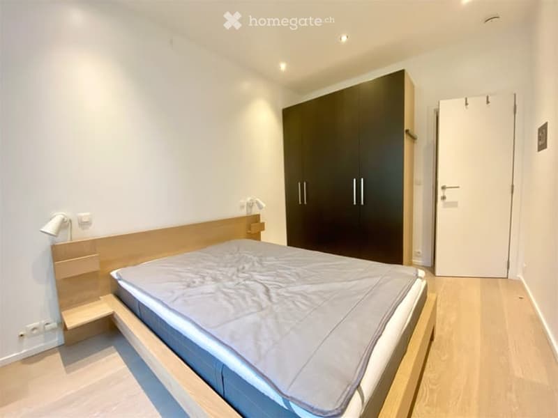 Modern bedroom with a wooden bed frame, grey bedding, a large dark wardrobe, and a wall-mounted lamp. The room features light wooden flooring and white walls.