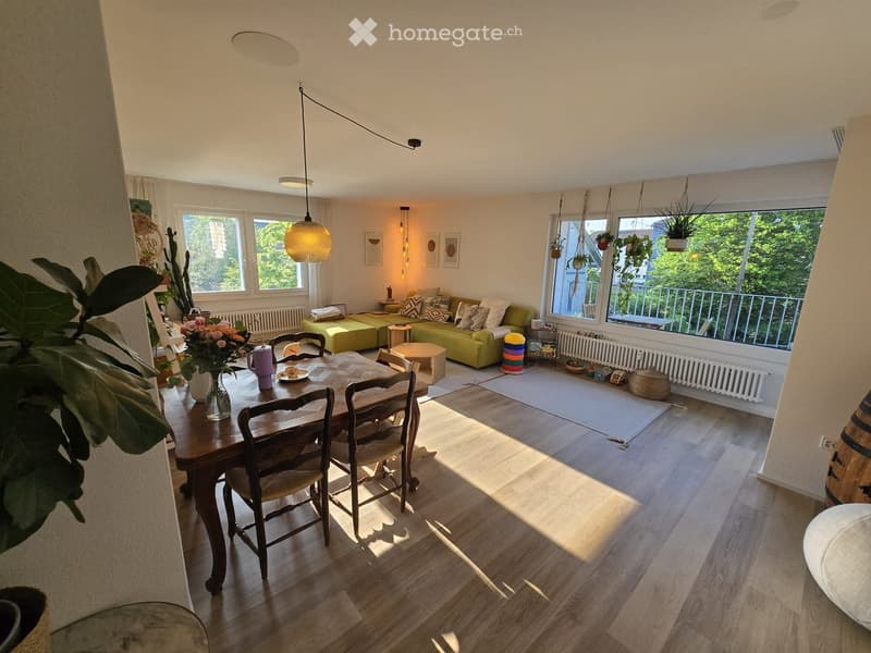 Bright living and dining area with wooden flooring, large windows, green sofa, dining table with chairs, pendant light, and decorative plants.