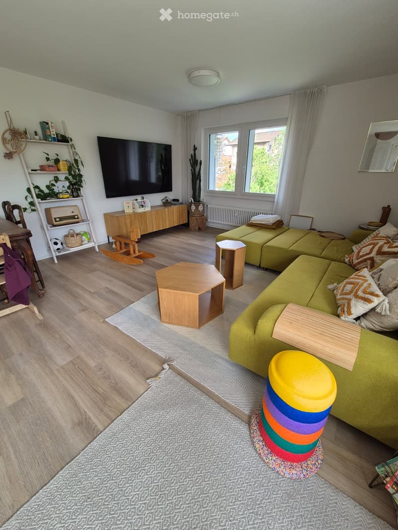 Modern living room with wooden flooring, large windows, green sofa, coffee table, TV, and decorative items.
