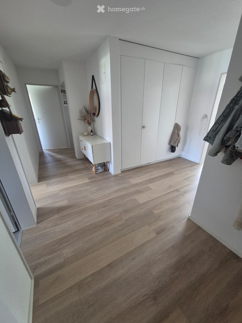 Bright hallway with wooden flooring, white walls, built-in wardrobe, small console table with decorative items, and coat rack with jackets.