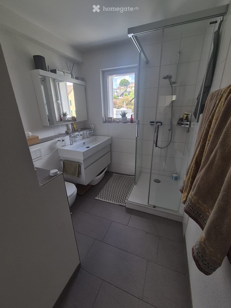 Modern bathroom with a white sink, mirror, toilet, and a glass-enclosed shower. The room features tiled walls and floor, a towel hanging on the side, and a small window offering a view of the outside.
