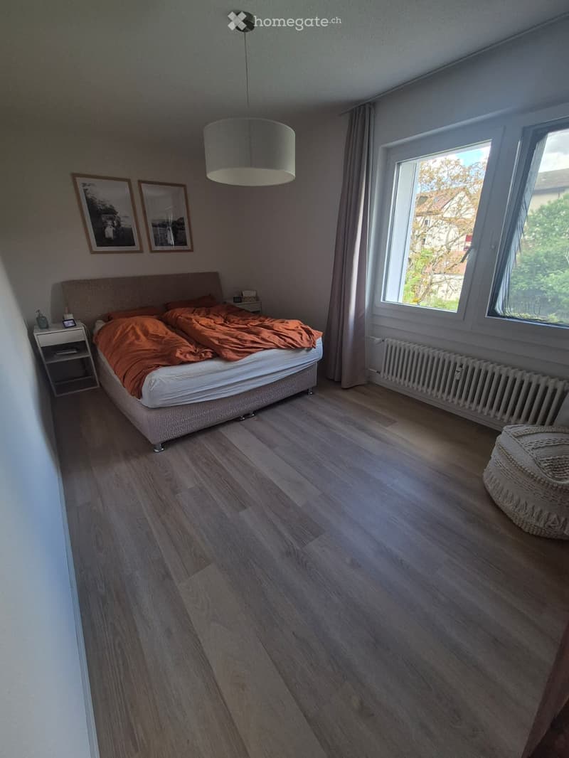 Bedroom with wooden flooring, large window with curtains, bed with orange bedding, nightstand, lamp, radiator, and picture frames on the wall.