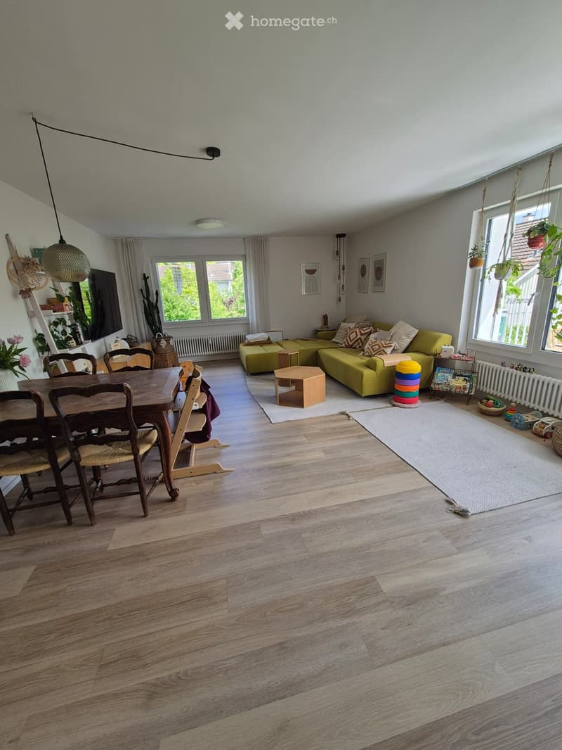 Bright living room with wooden flooring, large windows, green sofa, dining table with chairs, small wooden stool, and decorative items including a mirror and plants.