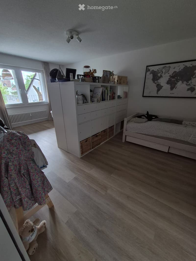 Bright bedroom with wooden flooring, large window with curtains, bed with white bedding, storage unit with books and toys, world map on wall, and a small stool with a floral blanket draped over it.