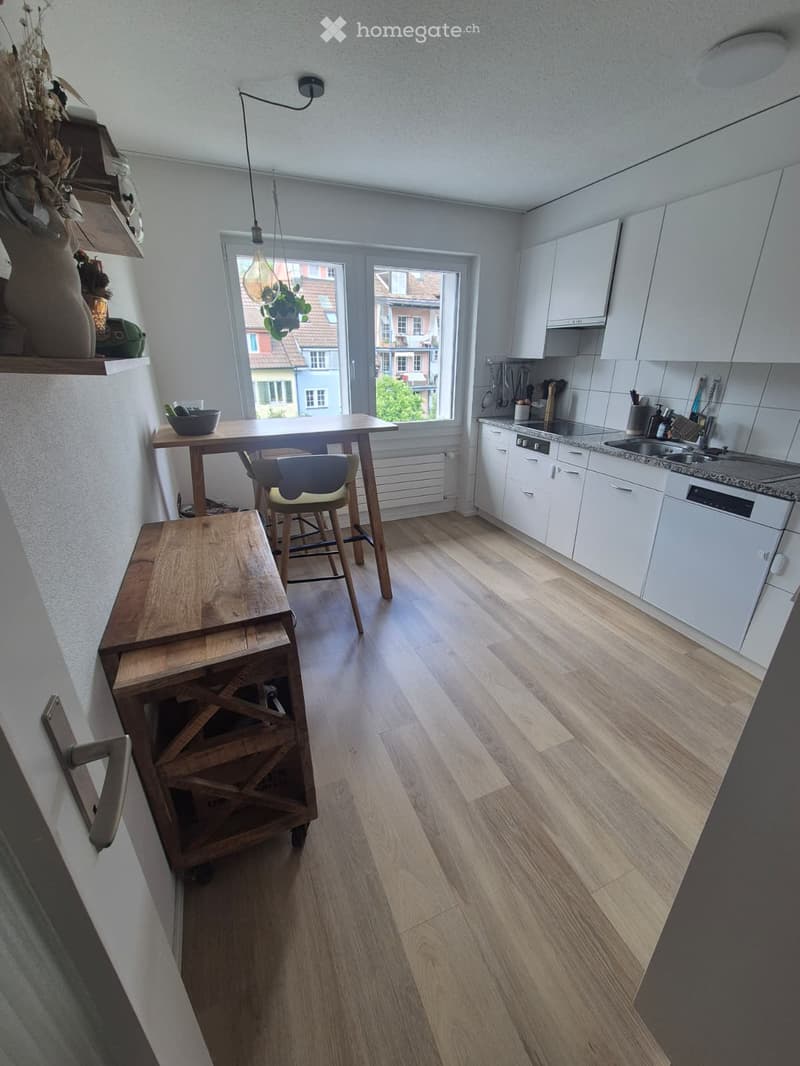 Modern kitchen with white cabinets, wooden flooring, and a dining area with a table and chairs. Features a sink, dishwasher, and various kitchen utensils.