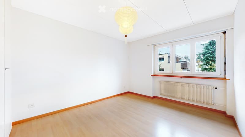 Empty room with white walls, wooden flooring, and a large window with a view of greenery. Features a radiator below the window and a hanging light fixture.