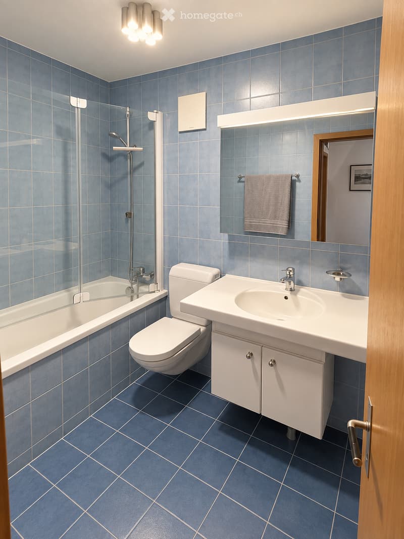 Modern bathroom with blue tiles, white fixtures, bathtub, toilet, sink, mirror, and towel.