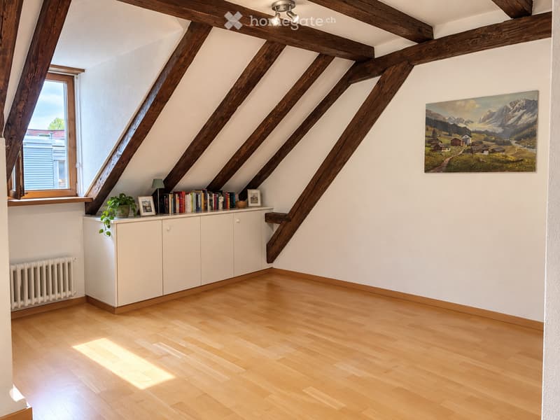 Bright attic room with wooden beams, white walls, wooden floor, large window, cabinet with books and a plant, and a painting on the wall.