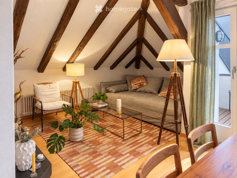 Cozy living room with exposed wooden beams, a sofa, armchair, coffee table, floor lamp, and decorative plants.