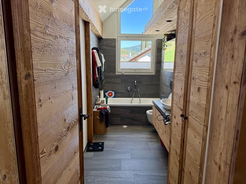 Modern bathroom with wooden walls, a bathtub, sink, toilet, and a large window offering a scenic view.