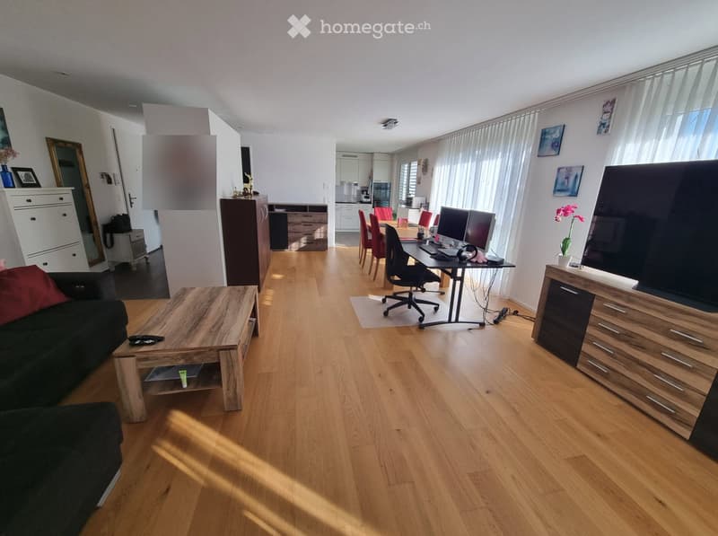 Open-plan living and working space with wooden flooring, large windows, sofa, coffee table, desk, chairs, and TV stand.