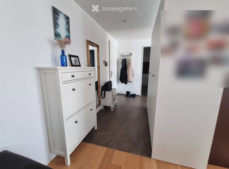A modern hallway with white walls, wooden flooring, and a white dresser with drawers. A mirror, vase with flowers, and framed picture are on top of the dresser. A coat rack with jackets and a small table with a bag are visible further down the hallway.