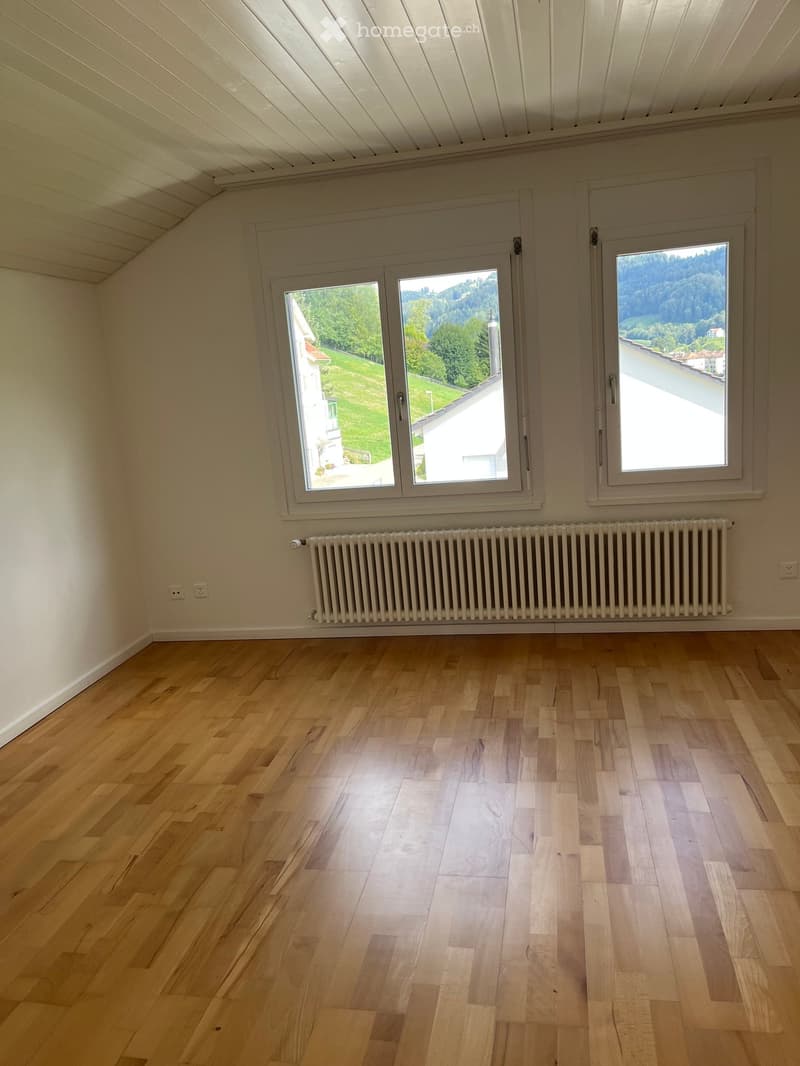 Empty room with wooden flooring, white walls, large windows, radiator, and visible electrical outlets.