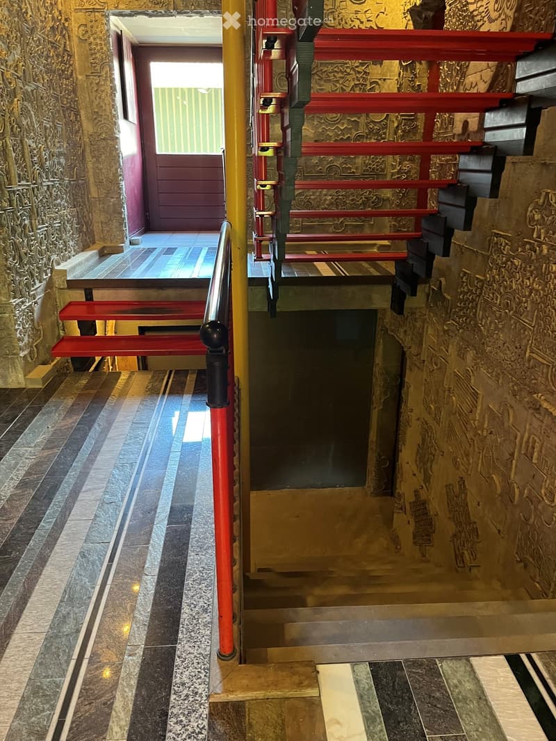 Staircase with red handrails and ornate stone walls, leading to a doorway with a window above it.
