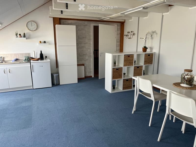 A modern, open-plan kitchen and dining area with white cabinets, a blue carpet, and a white dining table with chairs. The kitchen features a refrigerator, sink, and various small appliances.