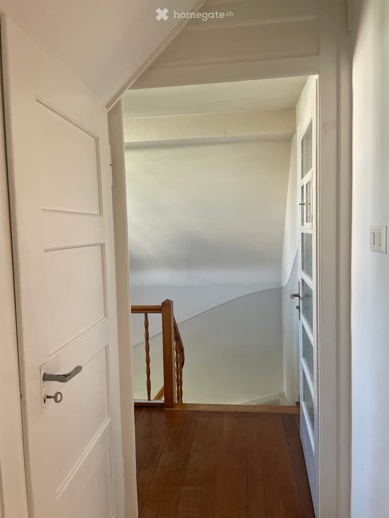 A narrow hallway with wooden flooring, white walls, and a staircase leading upwards. The hallway features a white door on the left and a small window on the right wall.