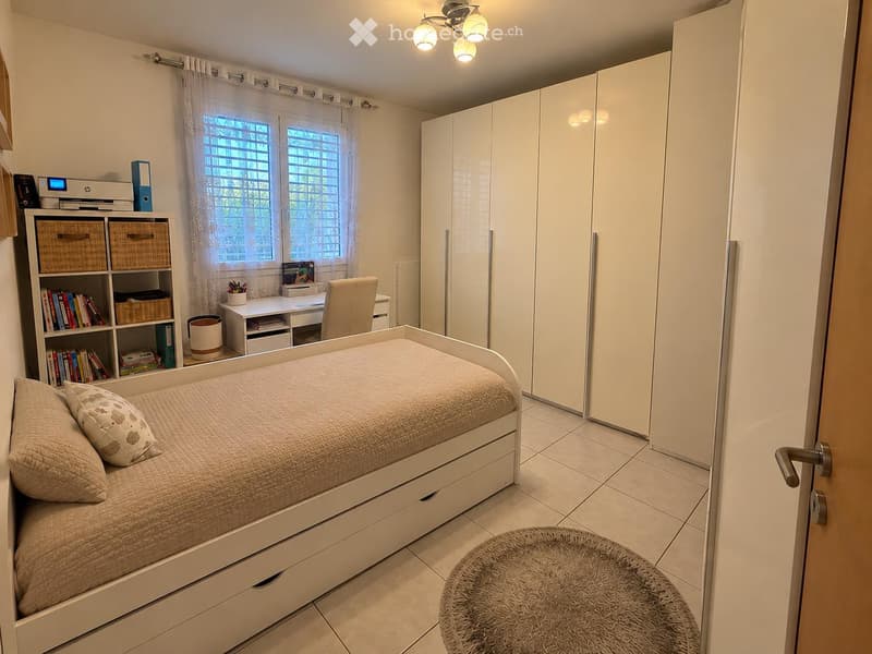 A modern, well-lit bedroom with a single bed, a desk, a bookshelf, and a large wardrobe. The room features a large window with curtains and a round rug on the floor.