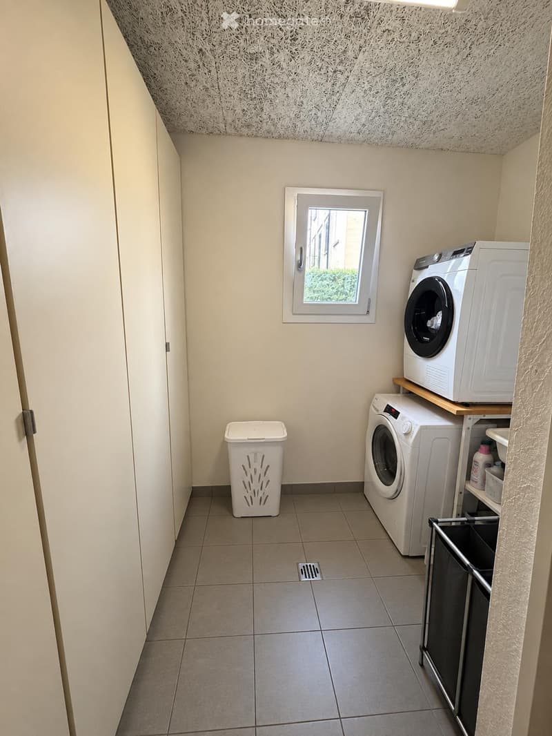 Small laundry room with tiled floor, white walls, a window, a washing machine, a dryer, a trash bin, and a small shelf with cleaning supplies.