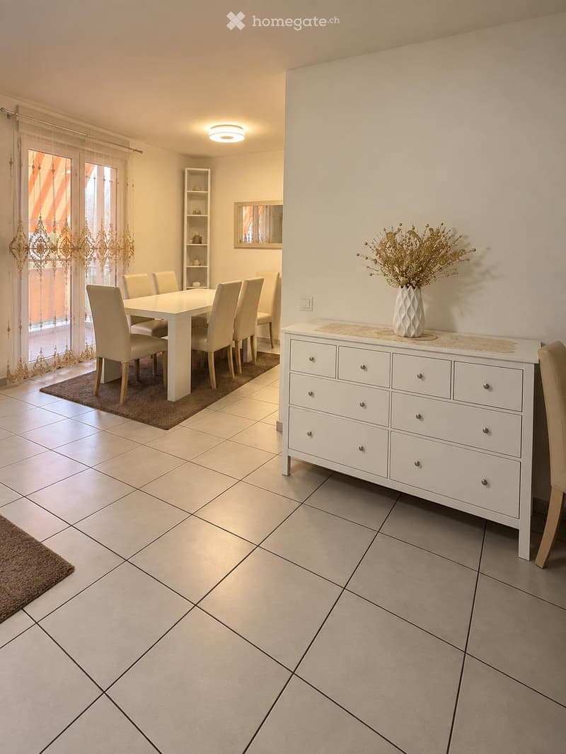 Dining area with a white dining table, six chairs, a white cabinet with a vase on top, and a mirror on the wall. The room has tiled flooring and large windows with curtains.