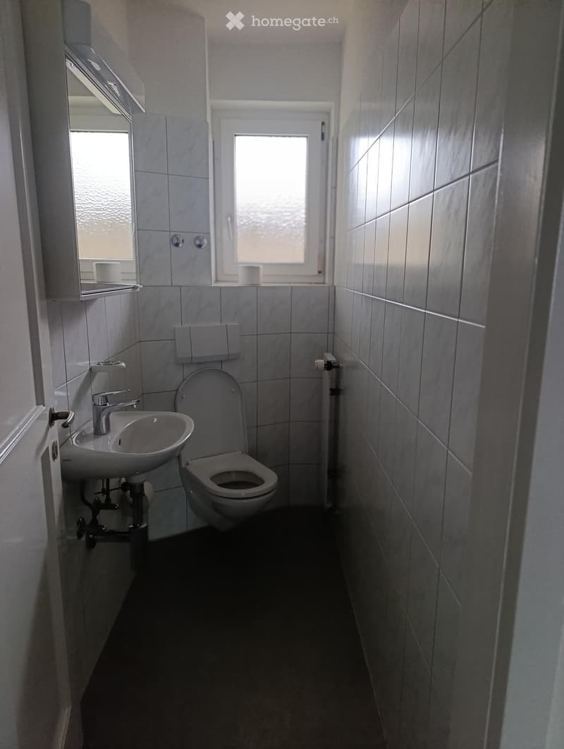Small bathroom with a toilet, sink, and mirror. The walls are tiled in gray, and there is a window above the toilet.