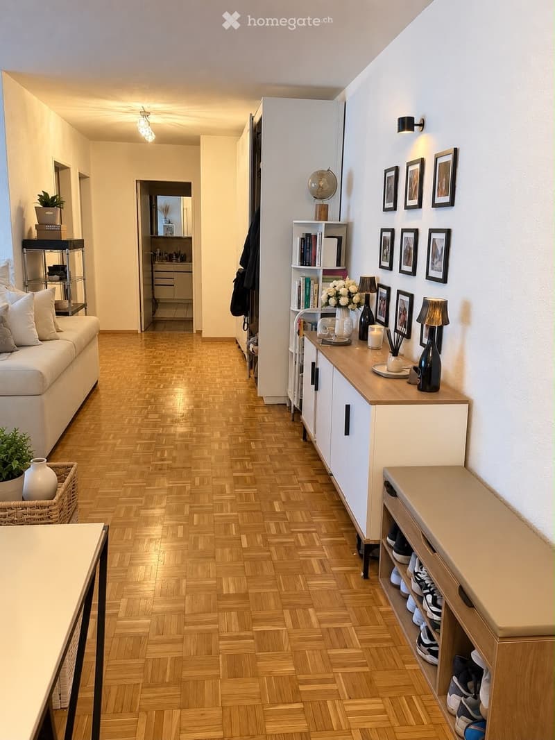 A well-lit hallway with wooden herringbone flooring, white walls, and modern decor including framed pictures, a small shelf with books and decorative items, and a shoe rack. The hallway leads to a living area with a white sofa and a kitchen area visible i