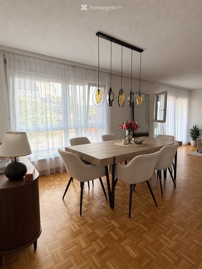 Modern dining room with a wooden dining table, white chairs, pendant lights, and a side table with a lamp and decorative items.