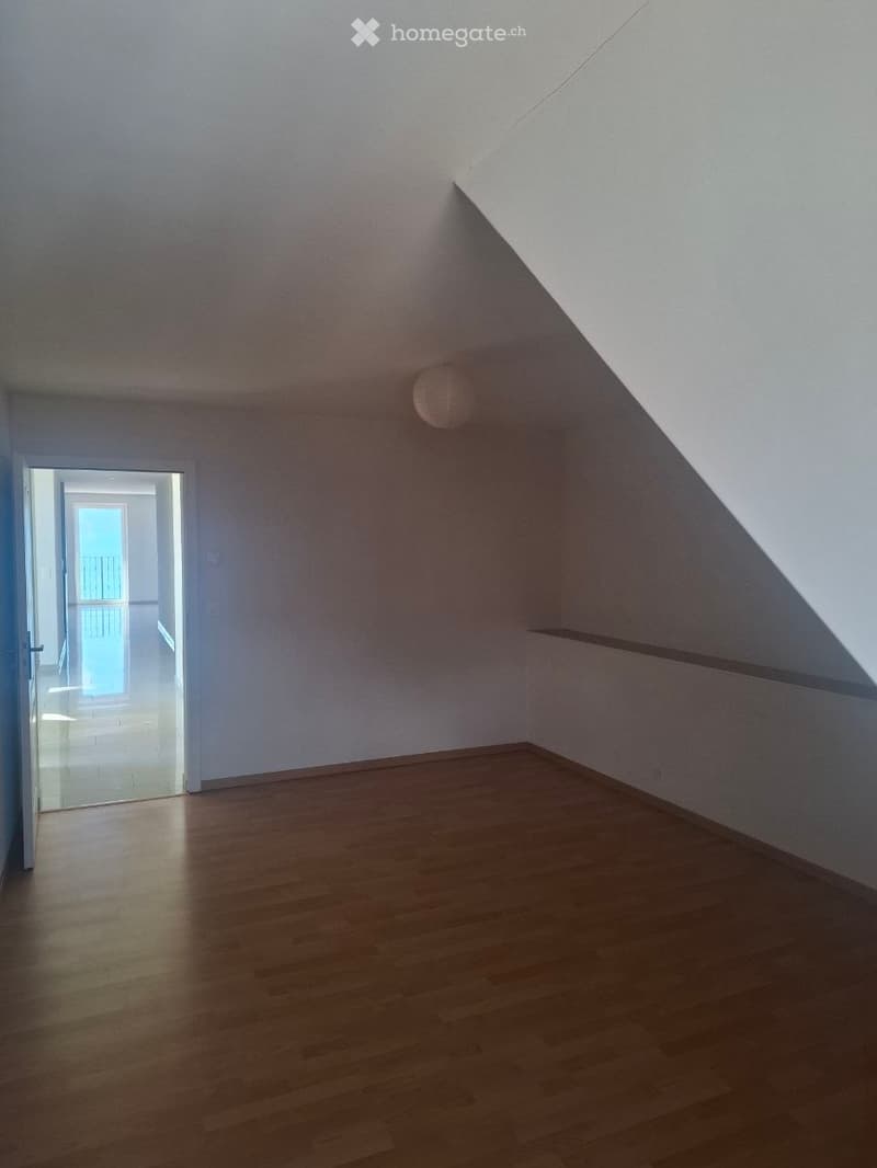 Empty attic room with wooden flooring, white walls, and a single light fixture. An open doorway leads to a balcony area with a glass door and visible outdoor space.